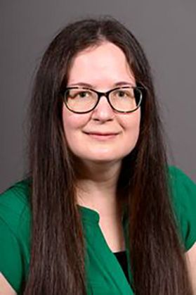 woman with very long dark hair year wears an emerald green blouse and dark plastic glasses. 