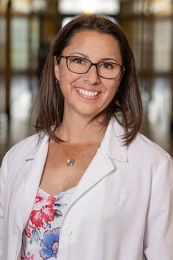 female with shoulder length dark hair and glasses wears a lab coat over a floral blouse