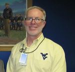 Male with grey hair and beard, and glasses wearing a light button down shirt with a WV logo embroidered on the chest. 