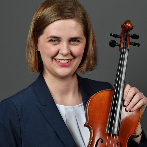 Female with chin length brown hair wears a blue blazer and white blouse. She holds her violin.