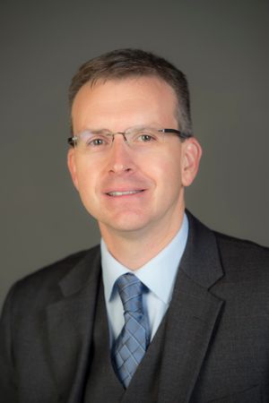 Male with short brown hair, rimless glasses wears a suit jacket and tie.
