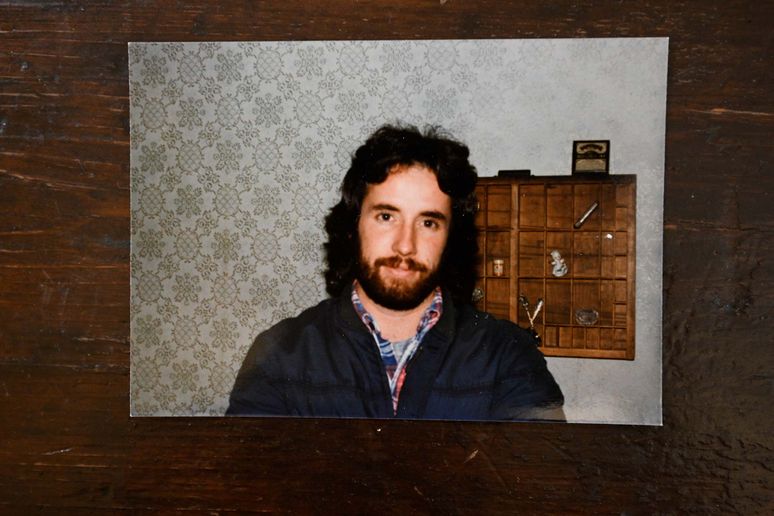 A younger James Nolan with a mullet, mustache and beard stands in front of a wallpapered wall