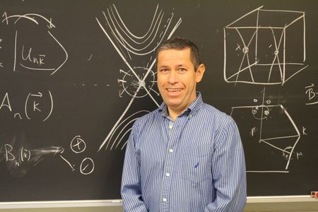 Aldo poses in front of a blackboard with physics problems. 