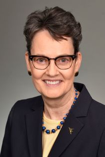 Woman with slightly wavy short hair and dark rimmed glasses wears black earrings and necklace and a sports jacket. 
