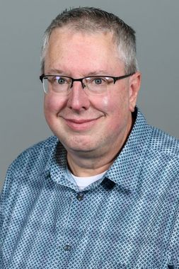 Male with short greying hair and wire rimmed glasses wears a plaid patterned button down shirt. 