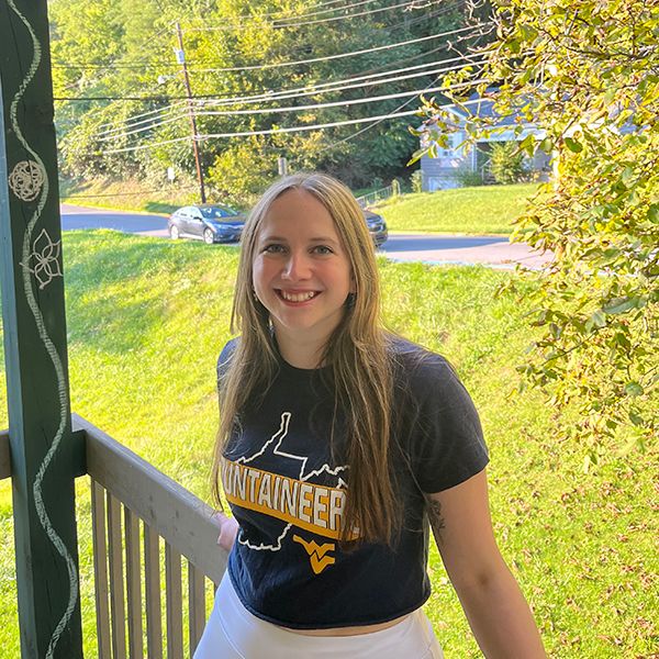 Female with long blonde hair and wearing a WVU Mountaineers t-shirt stands on her porch with grass and trees in the background.