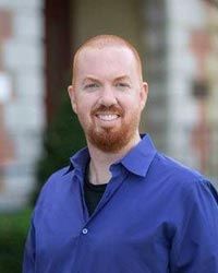 Man with a buzz cut, mustache and beard stands outside. He is wearing a blue button down shirt. 