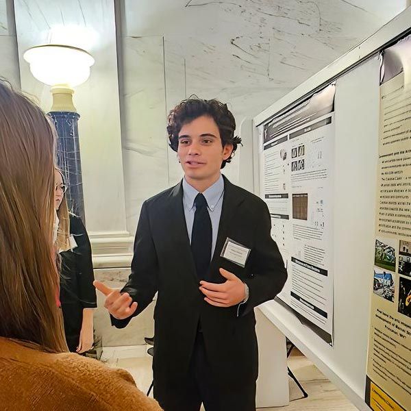 A student with short, curly dark hair stands in the rotunda of the State Capitol wearing a dark suit. To his left side is a research poster. A person with long brown hair stands with a back to the camera and listens.