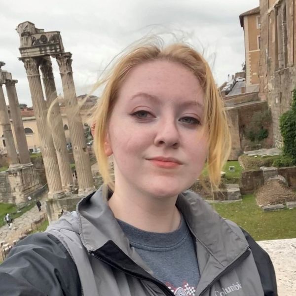 Headshot of WVU student Shelby Meador. She is pictured outside standing in front of ancient ruins. She is wearing a black and gray raincoat and has her blond hair pulled back. 