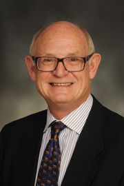 Man with grey hair and rectangular plastic frame glasses smiles for the camera. He wears a striped button down shirt, tie, and a dark suit jacket.