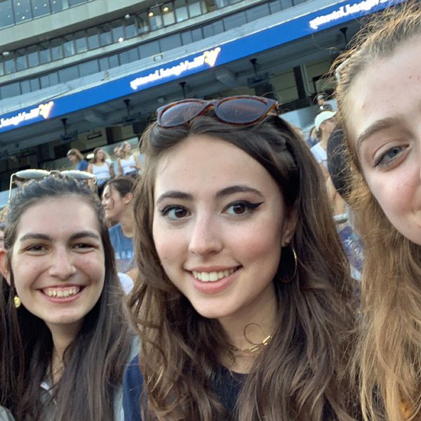 Female with long brown hair and sunglasses on her head poses with two friends while attending a WVU game. 