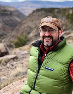 Man with beard and glasses wearing an outdoor puffer vest and a ballcap sits on a mountainside