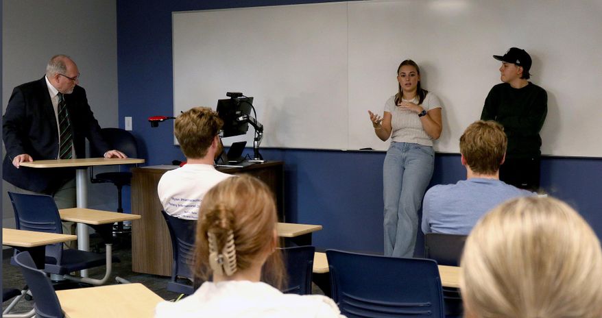 A male and a female student stand in front of a white board while giving feedback to a group of students seated in chairs. The Deal of Eberly College stands at a podium. 