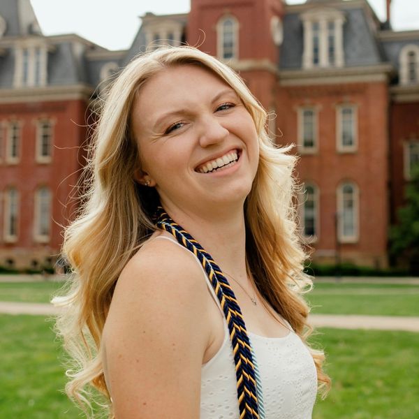 Headshot of WVU student Erin Langan. She is pictured outside in front of Woodburn Hall. She is wearing a white top with braided chords around her neck. She has long blonde hair. 
