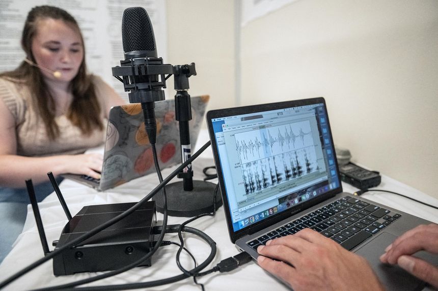 View of computer screen showing a linquistic analysis of speech patterns. Behind the laptop is a studio microphone and a girl in the background with a headset.