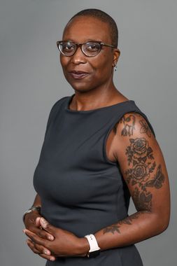A black woman with a shaved head wearing a black, sleeveless dress, cat eye glasses, an apple watch with white wristband, and small dangly earrings. She has monochrome rose tattoos on her left bicep. 