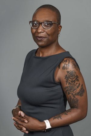 Black female with buzz cut hair and dark rimmed glasses wears a sleeveless dress to show her flower arm tattoos