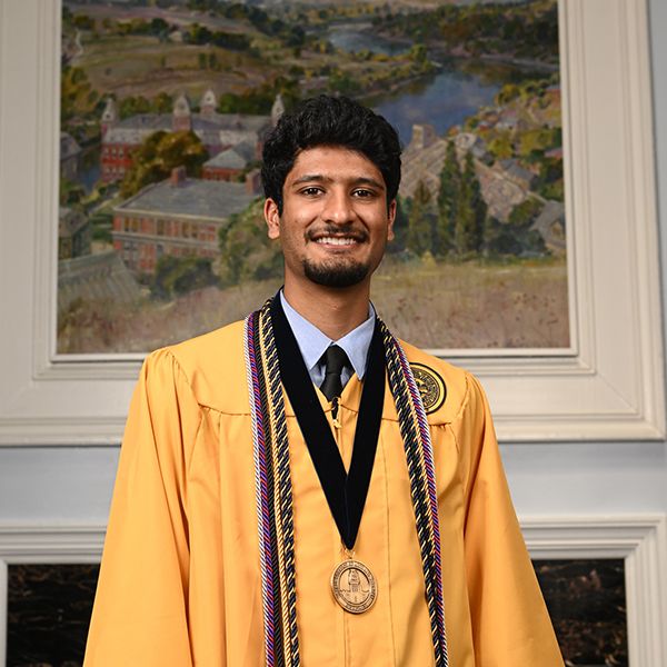 Male with dark hair, beard and mustache wearing a gold graduation robe, cords and medal poses in front of a picture of Woodburn Circle
