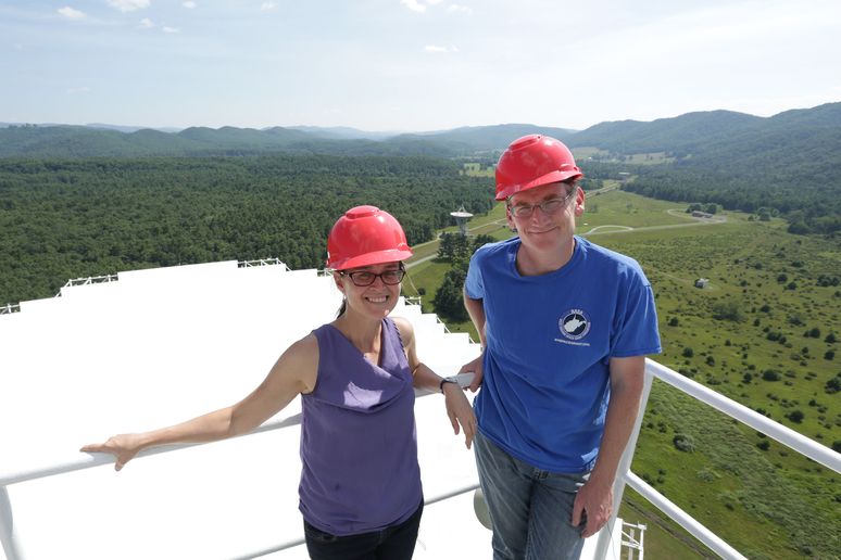 Male and female wearing red hard hats standing on a platform with West Virginia Mountains and the Greenbank radio telescope in the background.
