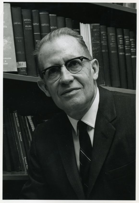 Black and white photo of Earl Core in front of a bookshelf wearing a suit jacket and tie.