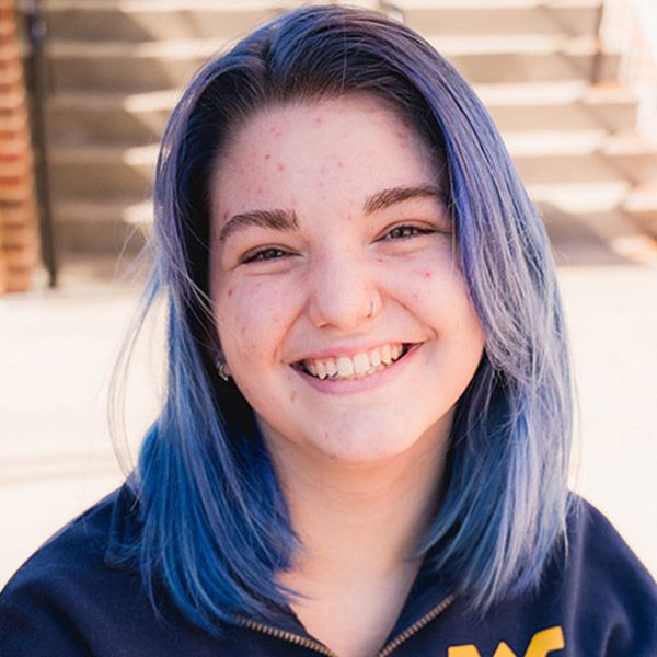 Girl with purple and blue faded hair wearing a blue shirt with gold WVU logo