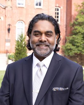 A brown skinned gentleman with salt and pepper beard and moustache. He has black wavy hair, is smiling, and wearing a suit and tie with a pocket square. 