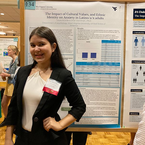 Female with long dark hair wears a black suit jacket and a white blouse poses in front of her research poster.