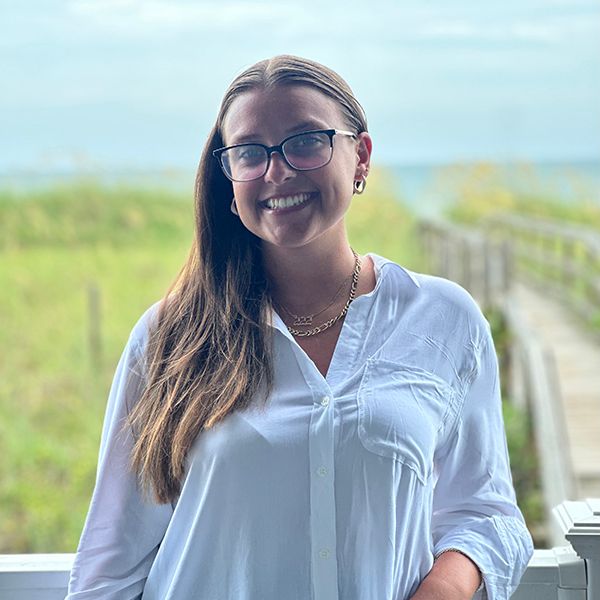 Female with long brown hair and glases wears a white shirt and a necklace. She stands in front of a boardwalk leading on an ocean.