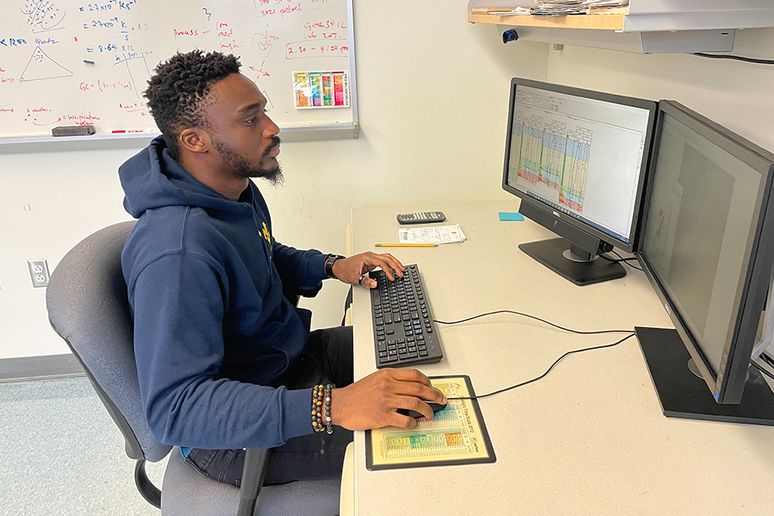Tobi Ore sits at a computer working with geology software. He is wearing a WVU hooded sweatshirt, has dark sin, dark curly hair, and a goatee. 