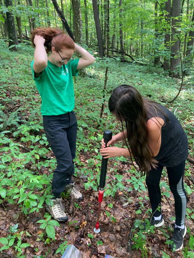 Female college student in t-shirt and jeans instructs a middle school girl with long hair and sleeveless shirt in out to take a soil sample.